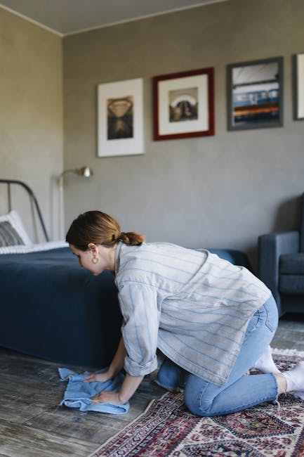 A person wearing a light-colored jumpsuit is operating a vacuum cleaner on a dark carpeted floor in a room with black wall panels, a radiator, and a speaker. The room is equipped with a large projecti
