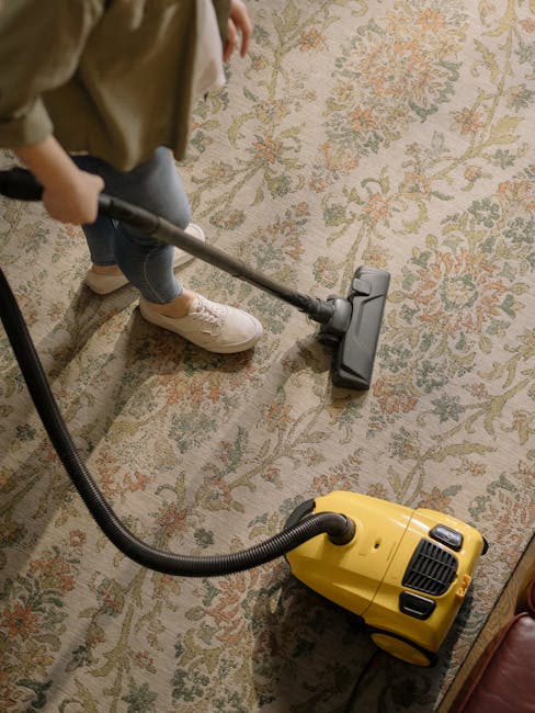 A person vacuuming a patterned area rug in a room with beige walls during a domestic cleaning session. The individual is using a yellow and black canister vacuum cleaner equipped with a flexible hose and a carpet cleaning attachment. The rug features intricate floral and foliage motifs in muted tones of green, orange, and cream, and appears clean and well-maintained. The room has soft, natural lighting that highlights the surface cleanliness and the textured surface of the rug. Hackney Carpet Cleaners specializes in surface cleaning and deep cleaning techniques aimed at maintaining hygiene and appearance of domestic carpets and rugs in Mare Street flats in Hackney.