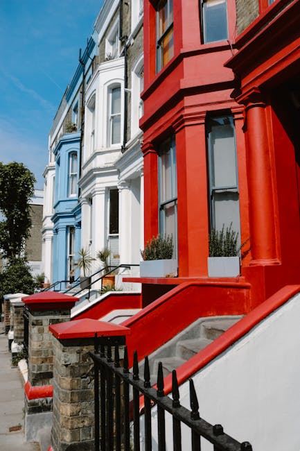 Exterior view of a row of Victorian terraced houses in London Fields E8, showcasing a mix of pastel blue, white, and bold red facades. The buildings feature large sash windows with black frames, some equipped with flower boxes filled with greenery. The steps leading to each entrance are made of concrete, with red-painted railings and black wrought-iron gates. The street is sunny with clear blue skies, and the sidewalk is lined with a few small plants and trees. The image emphasizes the architectural details and vibrant colors of the residential buildings, highlighting the characteristic London terraced street scene as seen from the sidewalk. The overall scene appears clean and well-maintained, with no visible dirt or debris, fitting the context of professional cleaning or maintenance of such properties that Hackney Carpet Cleaners might service.