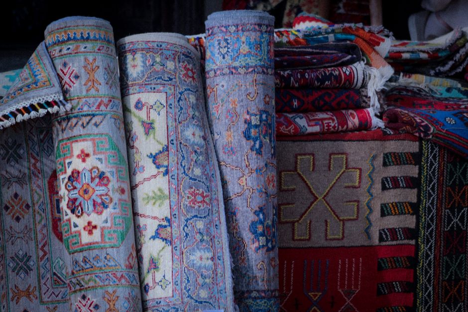 Close-up image of several rolled and folded traditional patterned rugs displayed vertically on a surface, with intricate designs in vibrant colors such as red, blue, green, and white. To the right, there is a neatly stacked pile of additional folded rugs with similar colorful patterns, resting on a flat surface, possibly a market stall or showroom. The background shows more rugs and textiles, suggesting a setting focused on rug sales or displays, with soft and even lighting highlighting the textures and detailed craftsmanship of the fabrics. The image emphasizes the variety and quality of oriental and modern rugs available for deep cleaning or maintenance services by Hackney Carpet Cleaners, aligning with surface cleaning and preservation practices for these textiles.