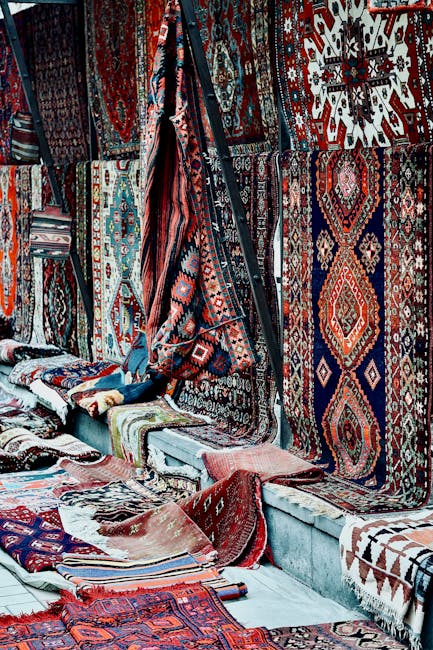 A display of numerous traditional Persian and oriental rugs hanging on a wall and laid out on the floor in a market setting, showcasing intricate patterns and vibrant colors such as red, blue, and white. The rugs feature geometric and floral designs, with some rolled or folded. Sunlight illuminates the scene, highlighting the textures and details of the woven surfaces. The setting is an indoor space with a white wooden platform and a concrete floor, with no visible cleaning tools or products in the image. This visual emphasizes the importance of surface cleaning and maintenance to preserve the quality and appearance of valuable rugs, as highlighted by Hackney Carpet Cleaners in their rug cleaning recommendations for Columbia Road market Hackney, Hackney.