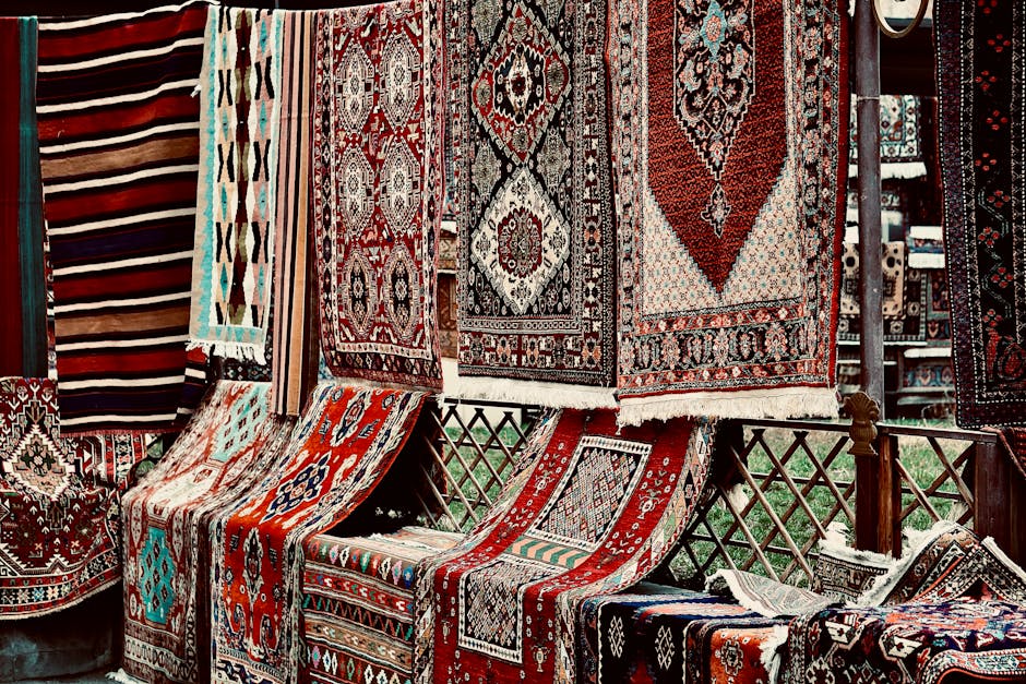 Close-up image of several rolled and folded traditional patterned rugs displayed vertically on a surface, with intricate designs in vibrant colors such as red, blue, green, and white. To the right, there is a neatly stacked pile of additional folded rugs with similar colorful patterns, resting on a flat surface, possibly a market stall or showroom. The background shows more rugs and textiles, suggesting a setting focused on rug sales or displays, with soft and even lighting highlighting the textures and detailed craftsmanship of the fabrics. The image emphasizes the variety and quality of oriental and modern rugs available for deep cleaning or maintenance services by Hackney Carpet Cleaners, aligning with surface cleaning and preservation practices for these textiles.