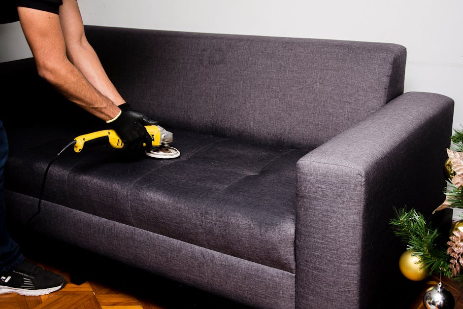 A person is performing surface cleaning on a dark gray fabric sofa with a handheld electric polisher, wearing black gloves and dark clothing, in a well-lit room. The sofa's fabric appears clean and slightly shiny, indicating recent deep cleaning work. To the right, there is a decorated Christmas tree with gold and pink ornaments, placed on a wooden floor, suggesting a residential setting. The room features a plain white wall background, and natural or artificial light highlights the sofa's surface, emphasizing its clean condition. This process is part of professional domestic cleaning services offered by Hackney Carpet Cleaners, focused on upholstery cleaning near Hackney Wick station, ensuring hygiene and freshness in home furnishings.
