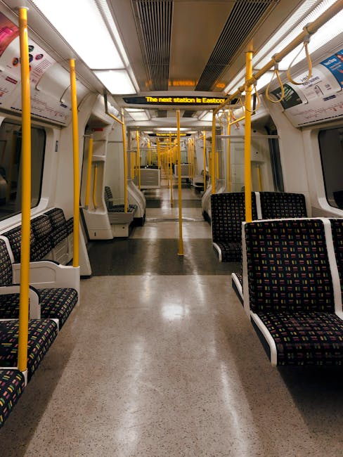 An empty interior of a modern train carriage featuring rows of upholstered seats with multicolored dotted patterns on dark backgrounds, arranged along both sides of the aisle. The floor is covered with smooth, beige vinyl flooring that appears clean and well-maintained. Bright white lighting illuminates the carriage evenly, reflecting off the clean surfaces. Bright yellow handrails run along the ceiling and sides, with ceiling-mounted handles hanging down for passenger support. The side windows are large, allowing natural light to enter and showing a blurred exterior scene. A digital display overhead shows the next station information. The overall setting is tidy, with no visible dirt or clutter, exemplifying professional cleaning and maintenance, similar to the deep cleaning services provided by Hackney Carpet Cleaners.
