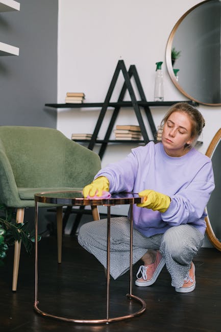 A person is performing surface cleaning on a dark gray fabric sofa with a handheld electric polisher, wearing black gloves and dark clothing, in a well-lit room. The sofa's fabric appears clean and slightly shiny, indicating recent deep cleaning work. To the right, there is a decorated Christmas tree with gold and pink ornaments, placed on a wooden floor, suggesting a residential setting. The room features a plain white wall background, and natural or artificial light highlights the sofa's surface, emphasizing its clean condition. This process is part of professional domestic cleaning services offered by Hackney Carpet Cleaners, focused on upholstery cleaning near Hackney Wick station, ensuring hygiene and freshness in home furnishings.
