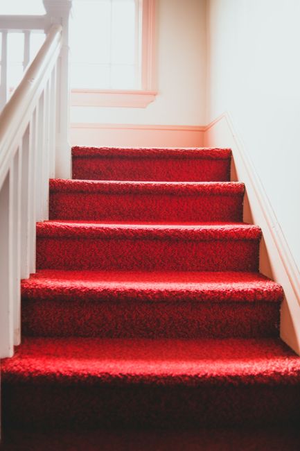 Close-up view of red carpeted narrow stairs in a room with soft lighting, highlighting the textured fabric surface that appears clean and well-maintained. The stairs are made of a plush, deep red mate